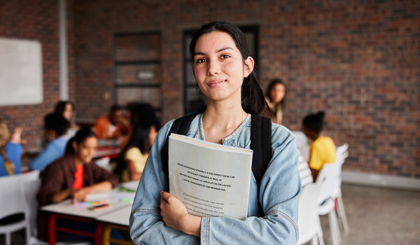 Young student smiling at the viewer, wearing a backpack and holding a stack of paper.